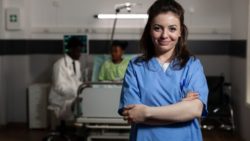 A nurse stands in a hospital room, with a doctor and patient in the background.